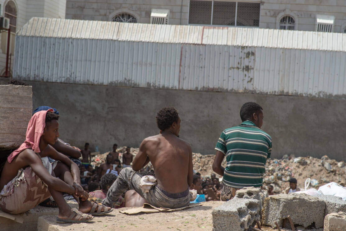 A group of male migrants sit together, looking at something outside of the camera's focus.