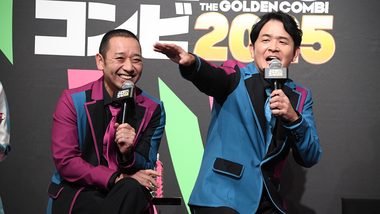 「THE GOLDEN COMBI 2005」と白い字が書かれた黒壁を背に、黒・青・紅色の衣装を着た2人がマイクを手に並んで座る。右側の男性は、右腕を左に伸ばしている。