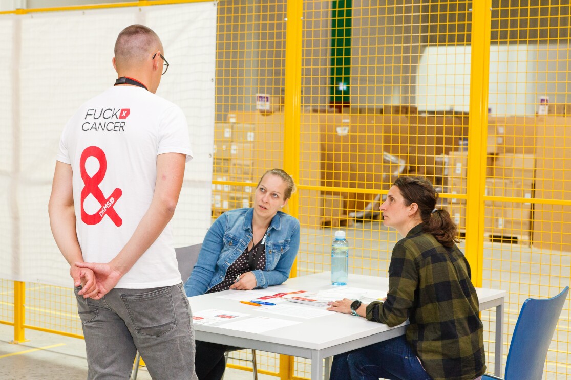 Ein Mann in Rückenansicht trägt ein T-Shirt mit der Aufschrift "Fuck Cancer", er steht in einem Logistikzentrum, vor ihm ein Tisch, an dem 2 Frauen sitzen