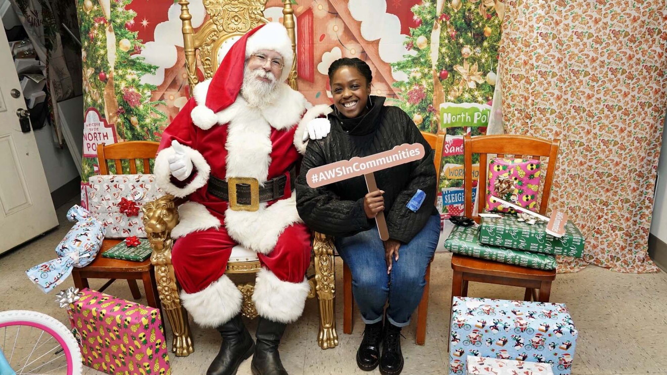 AWS in Communities: Santa Claus posing with smiling woman holding '#AWSInCommunities' sign