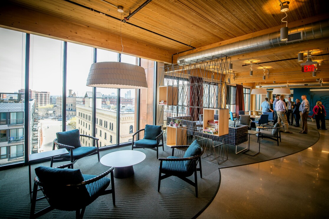 The view inside Amazon's Minneapolis tech hub - a wall of windows looks over nearby buildings, warm wood ceilings and exposed ventilation system are seen above casual co-working spaces. In the background, Amazonians gather.