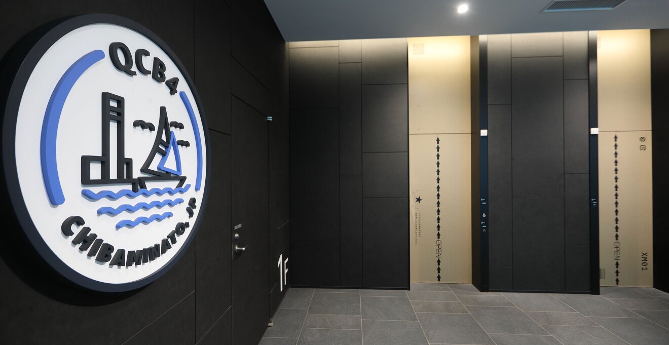 黒い壁のエレベーターホール。エレベーターのドアはダンボール箱を模したベージュ色。手前の壁にある円形看板には「QCBA4」、「Chibaminato. JP」の文字と、ヨットとタワーのアイコン。