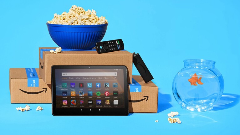 An image of two amazon boxes with a bowl of popcorn on top, a Fire TV stick and remote, and a tablet in front. Next to the boxes is a fishbowl and the background is bright light blue. 