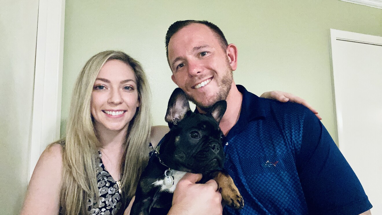 A man and a woman smile for a photo while holding a black French bulldog.