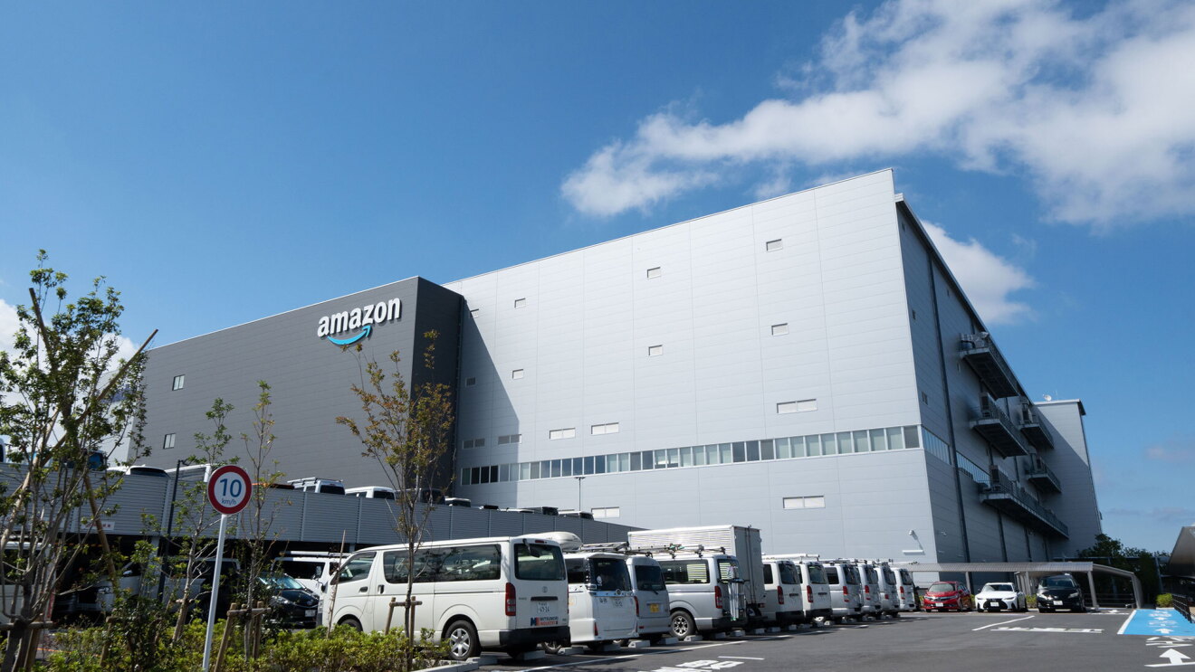 青い空をバックに建つ大きな建物。左側のダークグレーの壁にAmazonロゴ。右側の壁は薄いグレー。建物手間の立体駐車場に多数の車が停まっている。