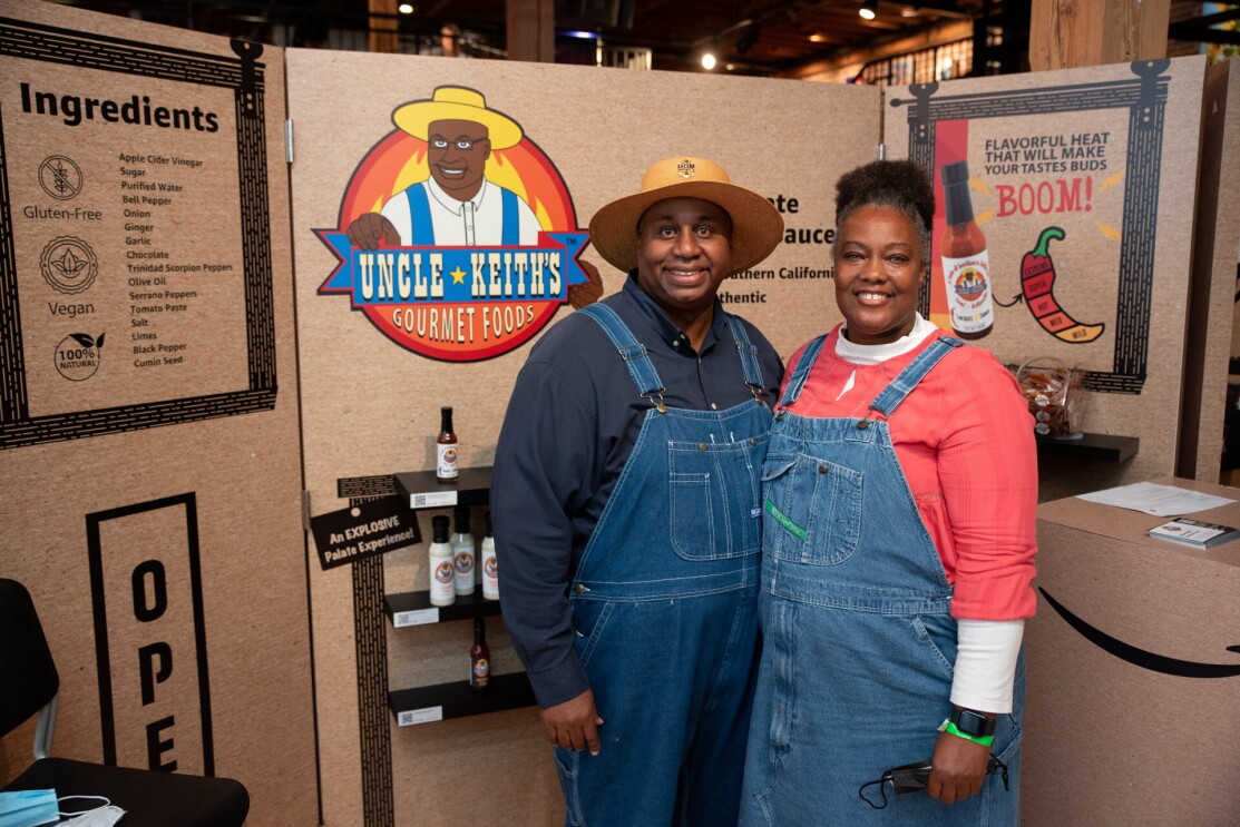 A man and a woman wearing overalls smile in front of sign that reads, "Uncle Keith's Gourmet Foods."