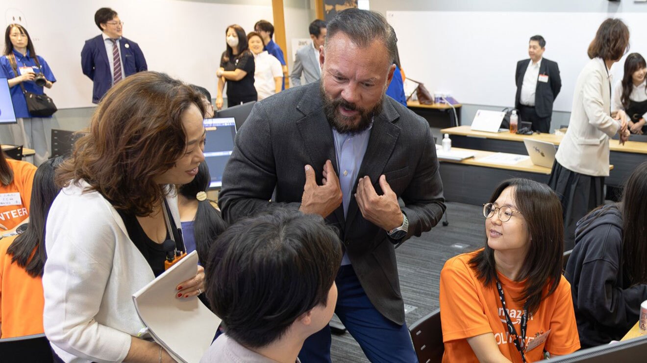 会議室のような空間で、椅子に腰かけた若者2人に、スーツ姿の男性が熱心に話しかけている。隣に白いジャケットの女性が立ち合い、背後には青いTシャツ着用のスタッフや参加者が見える。