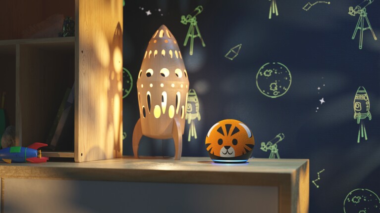 An image of an Echo Kids device next to a rocket shaped lamp sitting on a wooden table. The background is a dark blue wall with green space illustrations including a rocket, a planet and a telescope.