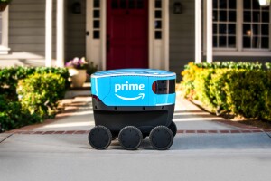 A bright blue, sleek, six-wheeled Amazon Scout delivery robot parked on the concrete walkway of an American home 