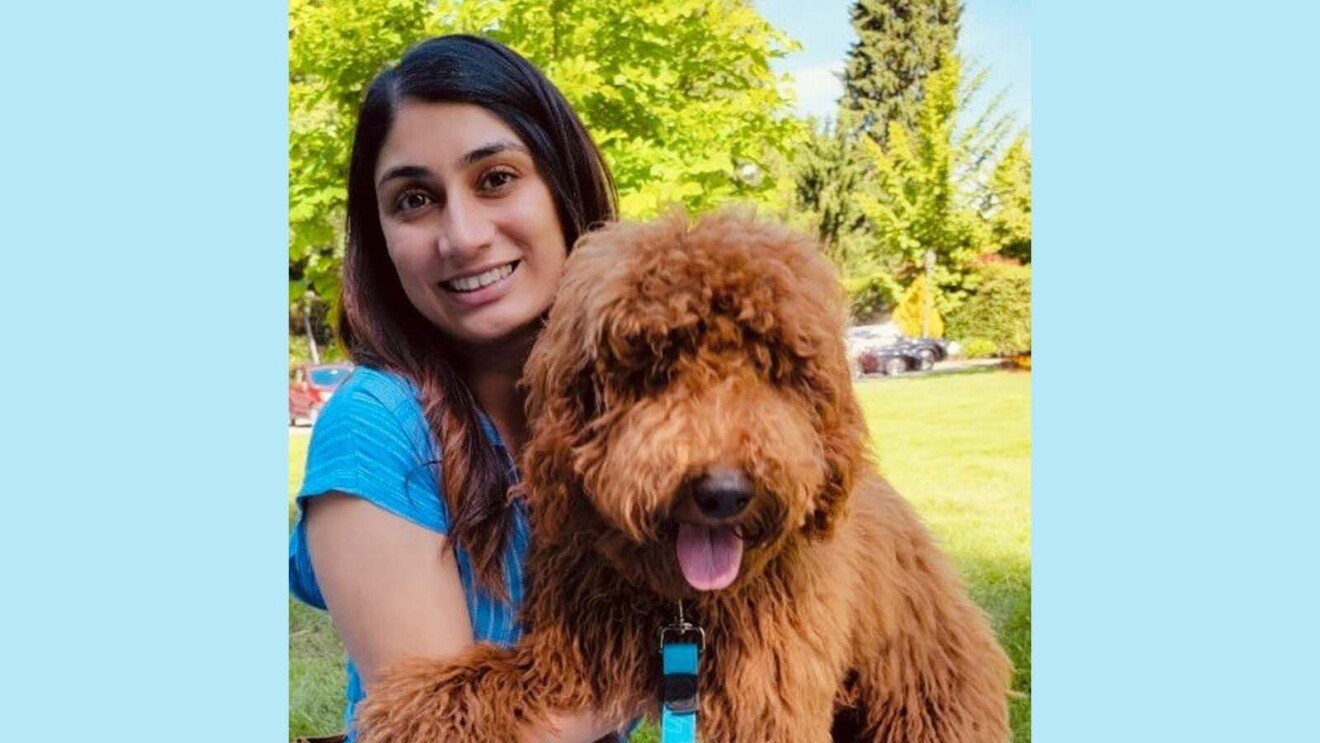 Poonam smiling and sitting on a blanket in a park while holding her mini goldendoodle, Clark.