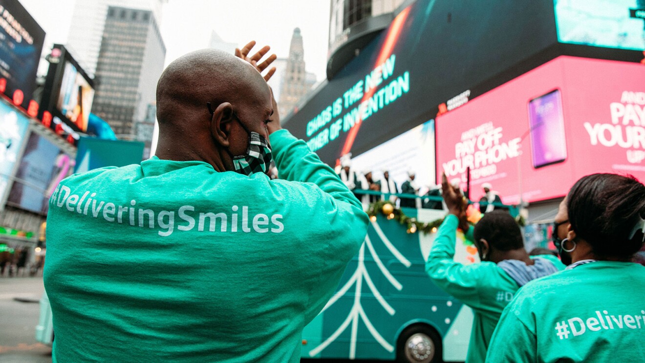 An image of a man clapping and smiling while wearing a mask and watching a choir perform on top of a double-decker bus.