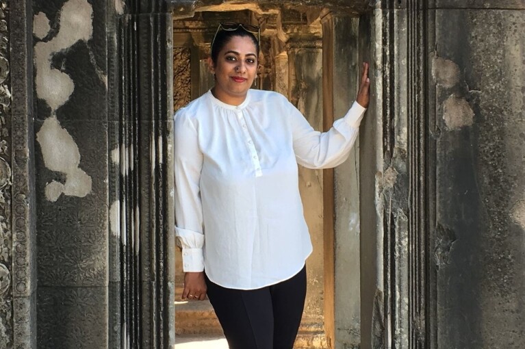 Gangamma stands between a doorway wearing a white blouse and sunglasses on her head.