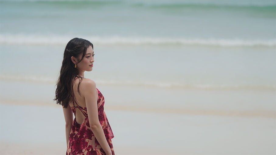 波が打ち寄せる浜辺に立つ女性。紅色の水着とビーチウェアを着用。