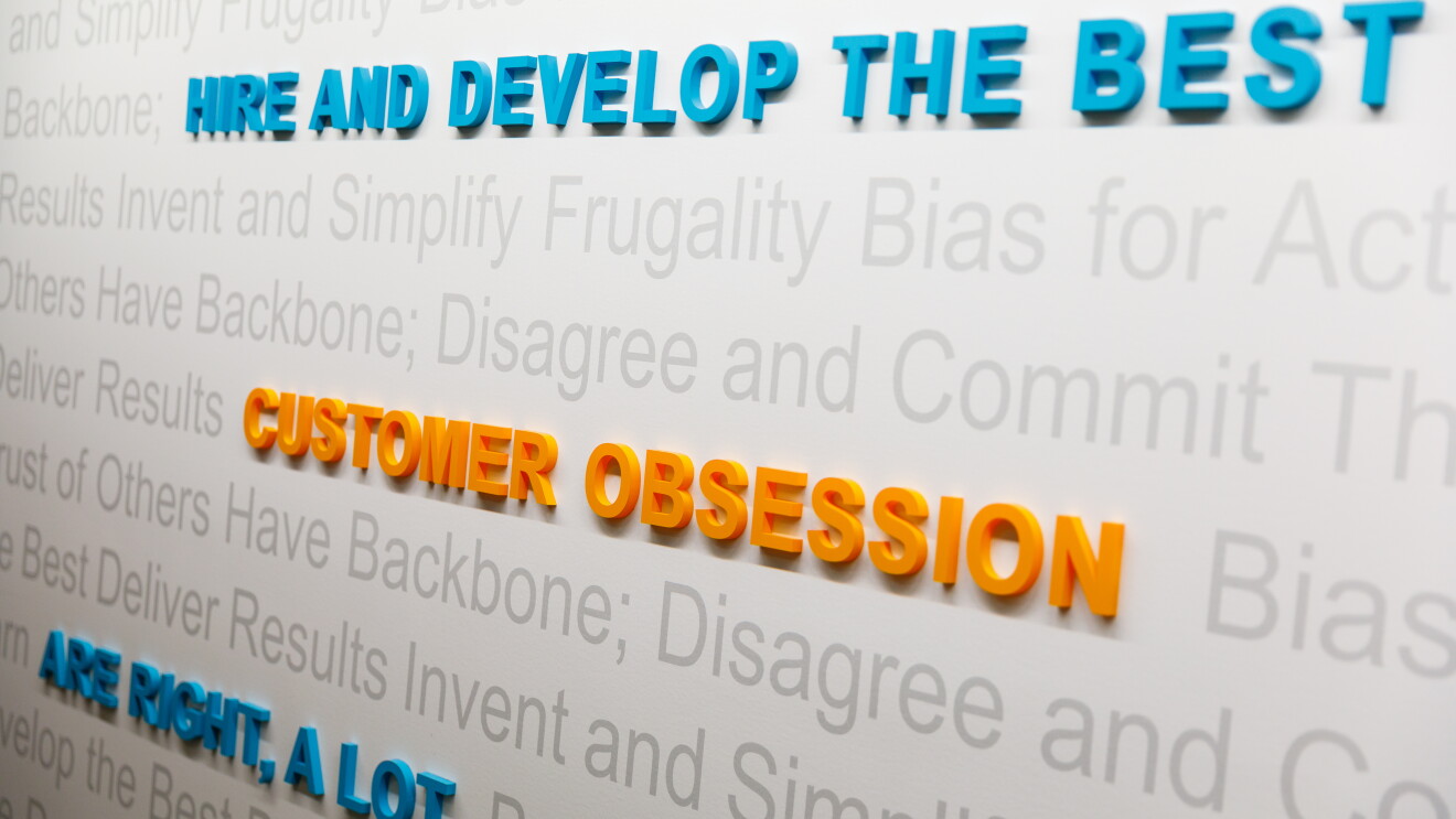 A visual display on a wall at Amazon, highlighting three of the leadership principles, with "customer obsession," "are right, a lot," and "hire and develop the best" in raised letters on a backdrop of other Leadership Principles.