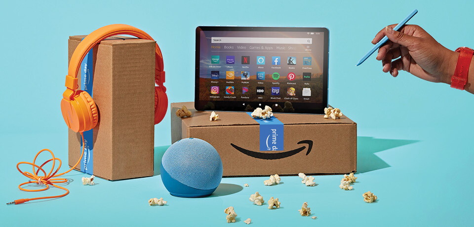 Image of an Amazon box with Prime Day tape, with a tablet on top of it, an Echo device in front of it, and another box with orange headphones.