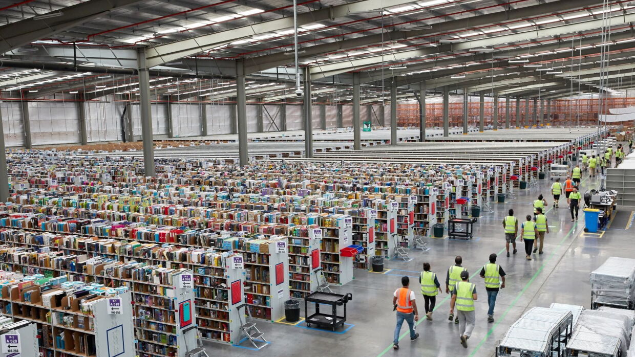 An image from the top of an Amazon fulfillment center. There are rows of shelves of products on the left of the photo and employees with their safety vests on walking alongside a walkway to the right of the photo.