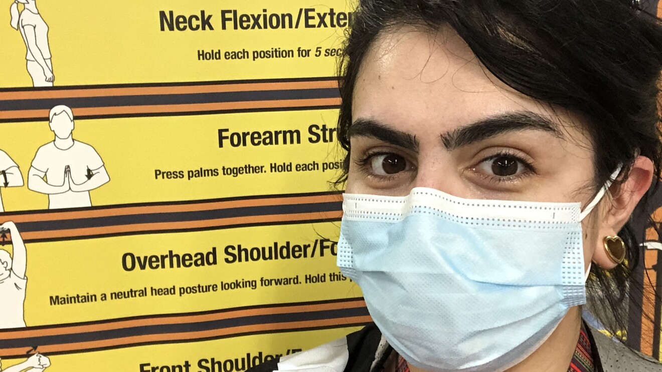 Beka Perez, an Amazon associate, smiles for a photo in front of a sign at an Amazon fulfillment center. The sign shows useful stretches for fulfillment center employees. Beka is wearing a face mask.