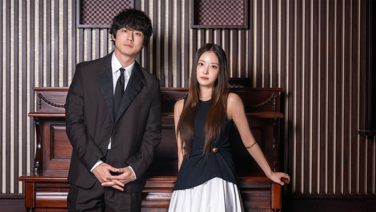 スーツを着ている男性（坂口健太郎さん）と黒いブラウスを着ている女性（イ・セヨンさん）は木材で作られたテーブルの前に立って並んでいる