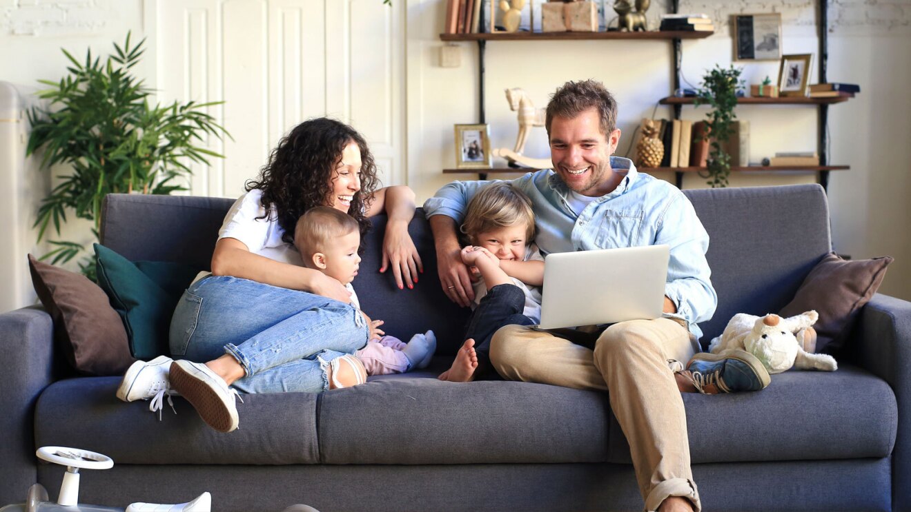 A family of four smiles and sits on a couch looking at a laptop.