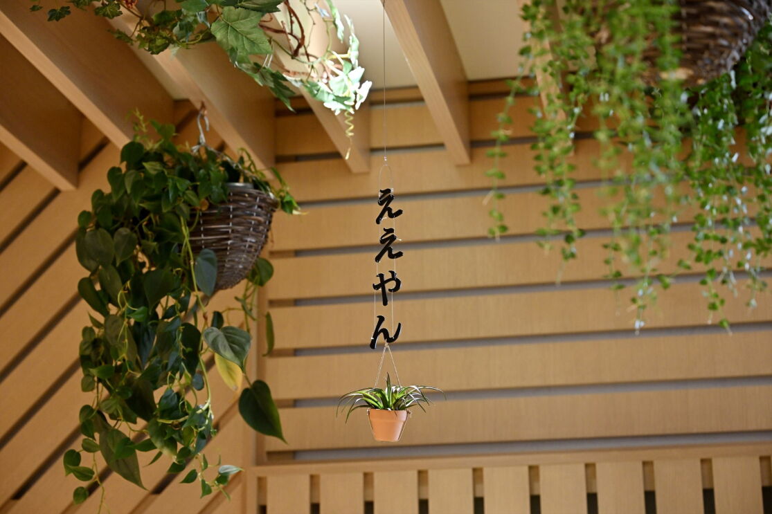 木材で作られた壁の前に縦書きの「ええやん」の文字が天井から吊り下げられている。その文字の下に小さな植木鉢に入った植物がある