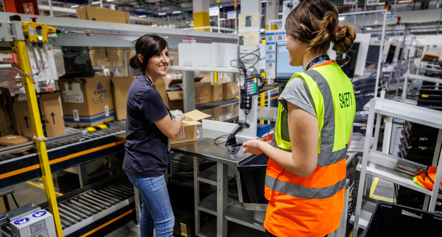 Ramona Moraru, supervisora de operaciones en la estación logística de Getafe. En el exterior del centro logístico con dos camiones de Amazon Prime de color azul con la sonrisa de Amazon en blanco. Ramona está hablando por un walkie talkie. Tiene el pelo claro, recogido en una cola, Con la mano derecha agarra el walkie talkie, tiene las uñas pintadas de negro. Lleva un tatuaje en la muñeca izquierda y un pulsera. Los ojos oscuros y maquillados y una mascarilla negra de Amazon. Lleva un chaleco de color naranja y un jersey negro con estrellas de color gris.  