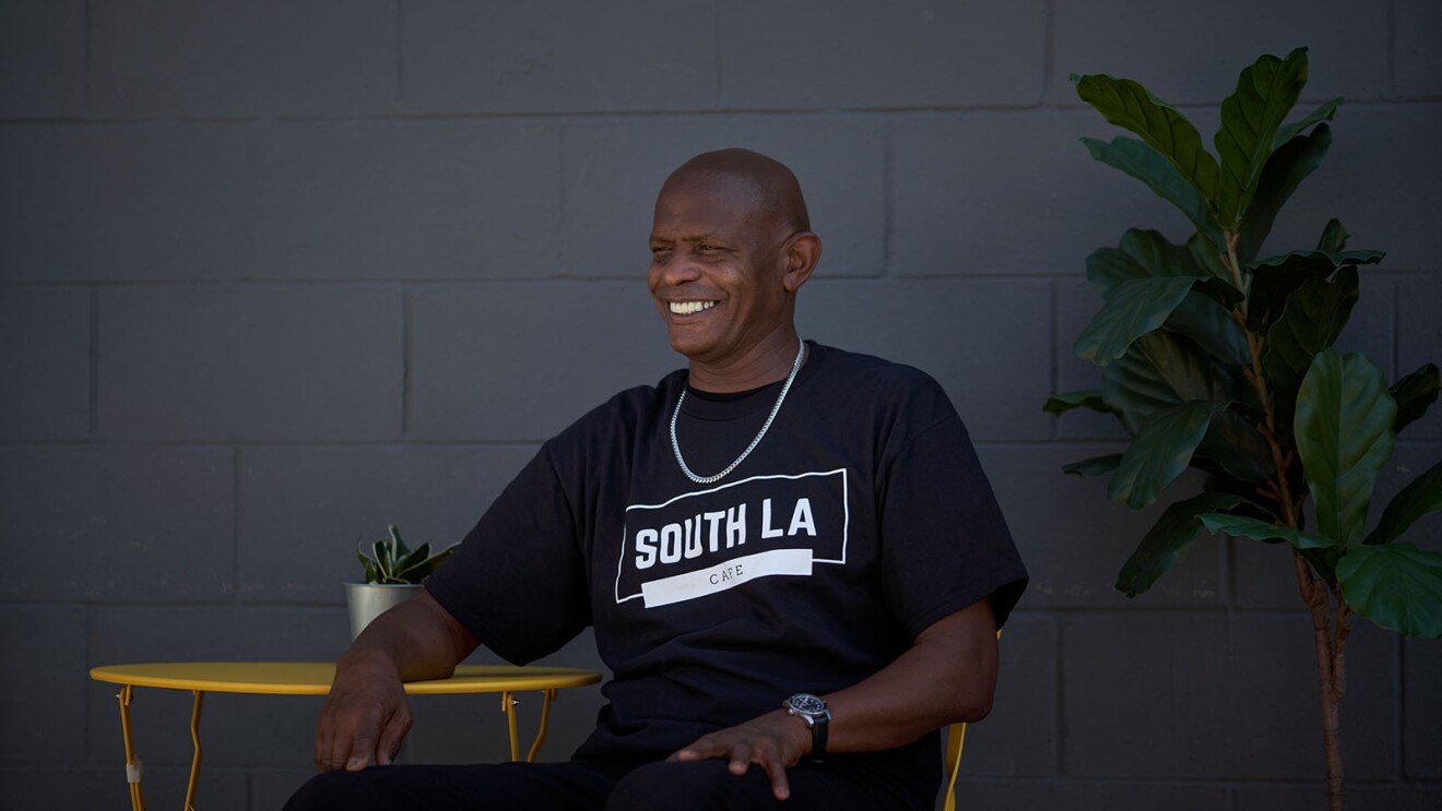 一个男人微笑着坐在一张黄色的户外桌子旁。他的衬衫上写着“南洛杉矶咖啡馆”