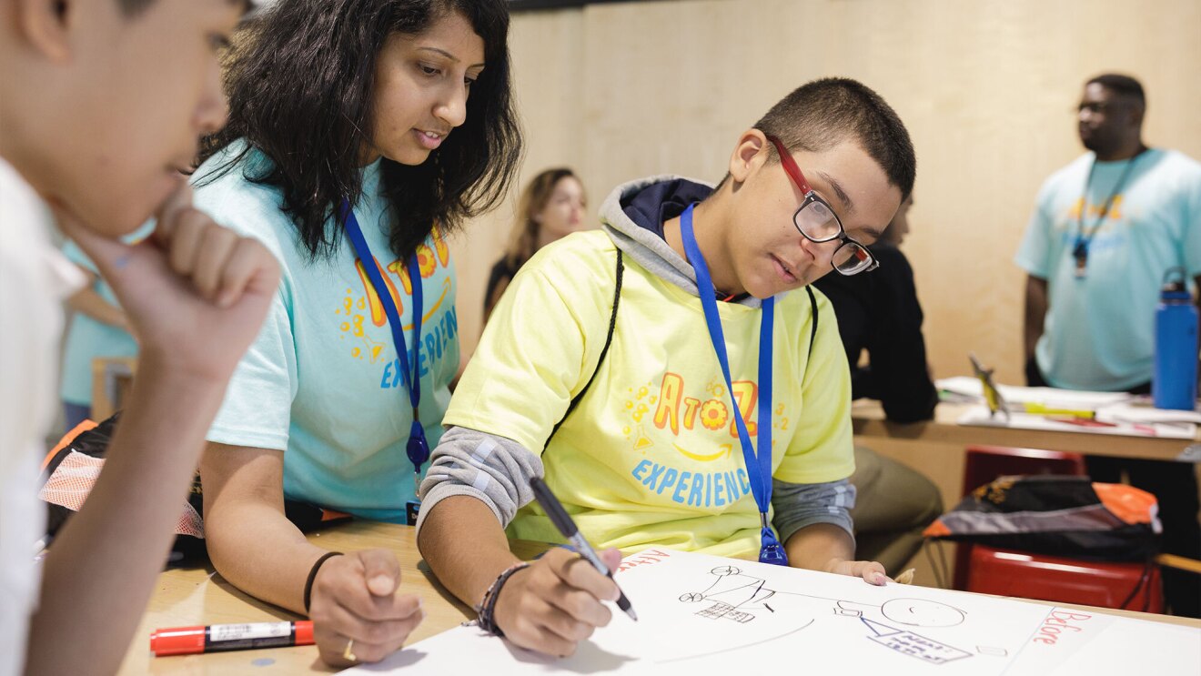 A student draws on a poster board with two instructors watching him.