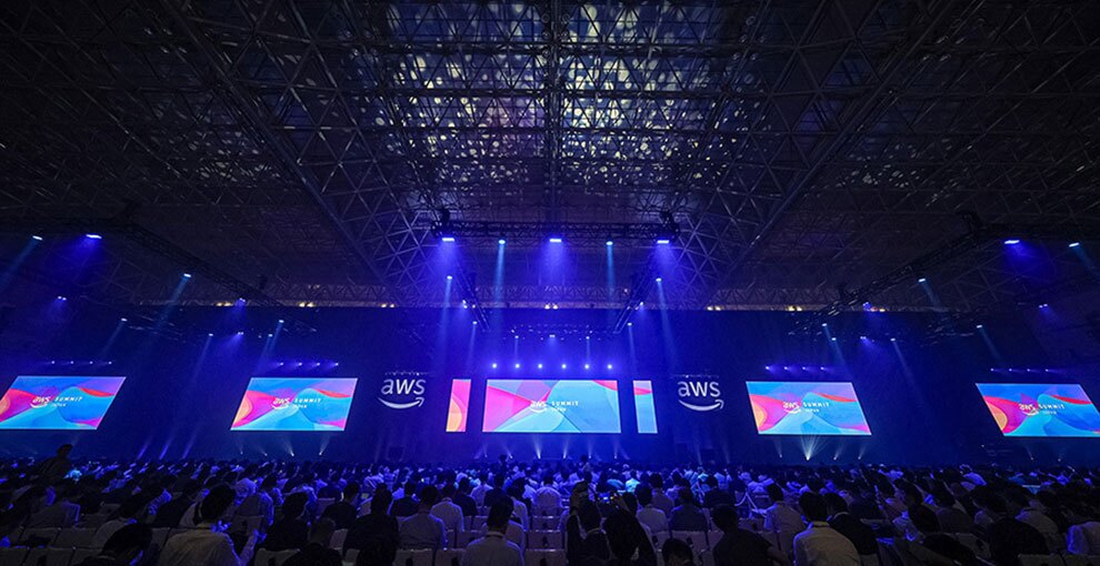 青いライトに照らされた薄暗い大型イベント会場。複数のスクリーンにAWSのロゴとスライドが映り、客席を聴衆が埋め尽くしている。
