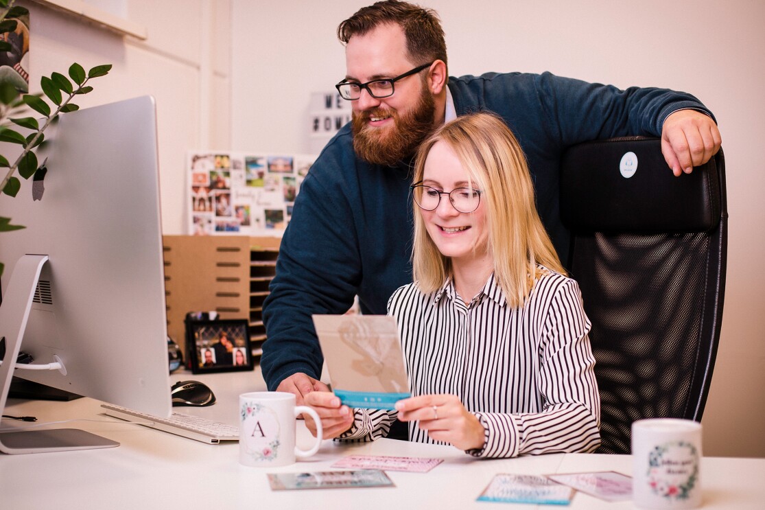 Eine Frau und ein Mann sitzen und stehen vor einem Computerbildschirm. Sie lächeln beide und schauen in den Bildschirm. Die blonde Frau hält eine Karte in den Händen, von der nur die Rückseite zu sehen ist. Sie befinden sich in einem Büro..