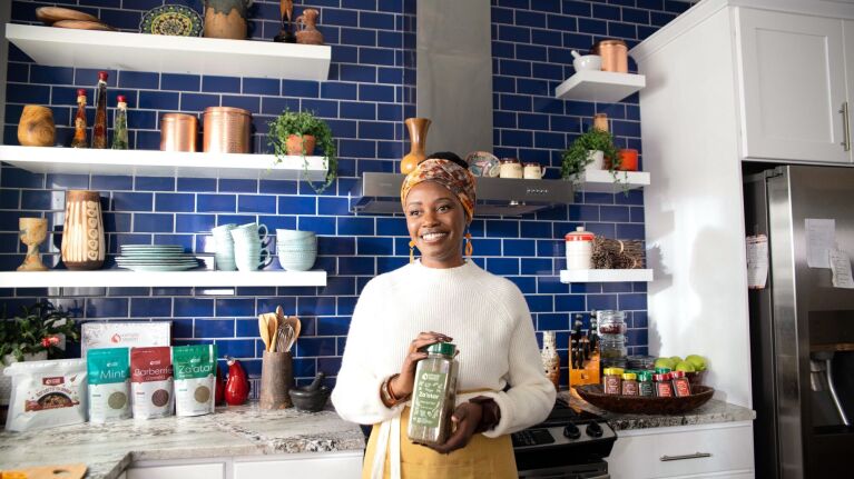 sylvia kapsandoy the founder of usimplyseason stands in front of a kitchen stove and countertop holding a carton of spice seasonings