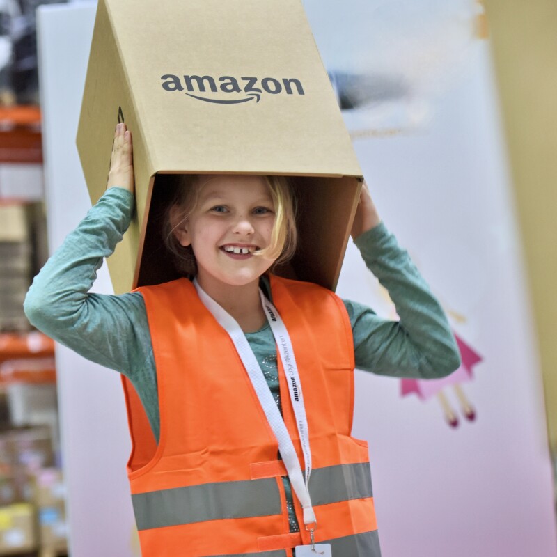 Ein kleines Mädchen in orangener Sicherheitsweste hält einen Karton mit Logo und Amazon Aufschrift hoch.