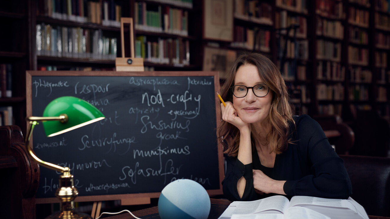 Susie Dent sitting at a table in a library next to a blackboard and with books and an Echo on the table. 