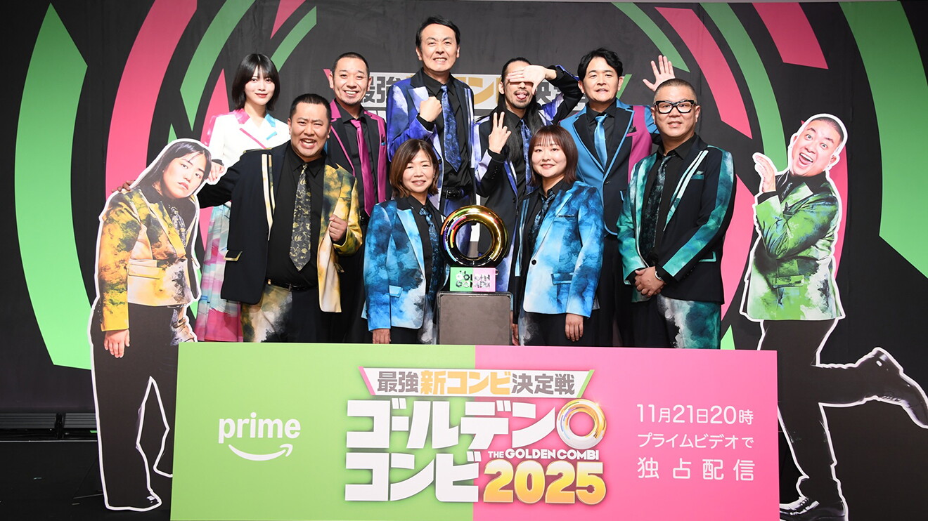 黒地にピンクと黄緑の曲線が入った背景に、派手な衣装を着た11人の人物が立ってカメラを見ている。中央に金色のトロフィー、脚の前にピンクと黄緑の横長パネル。