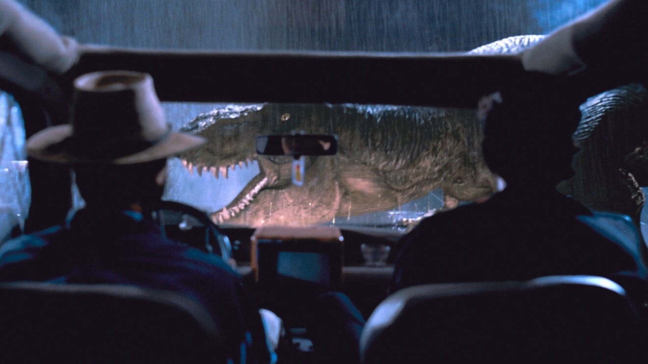 Tyrannosaurus rex looming over car windshield in rainy night.