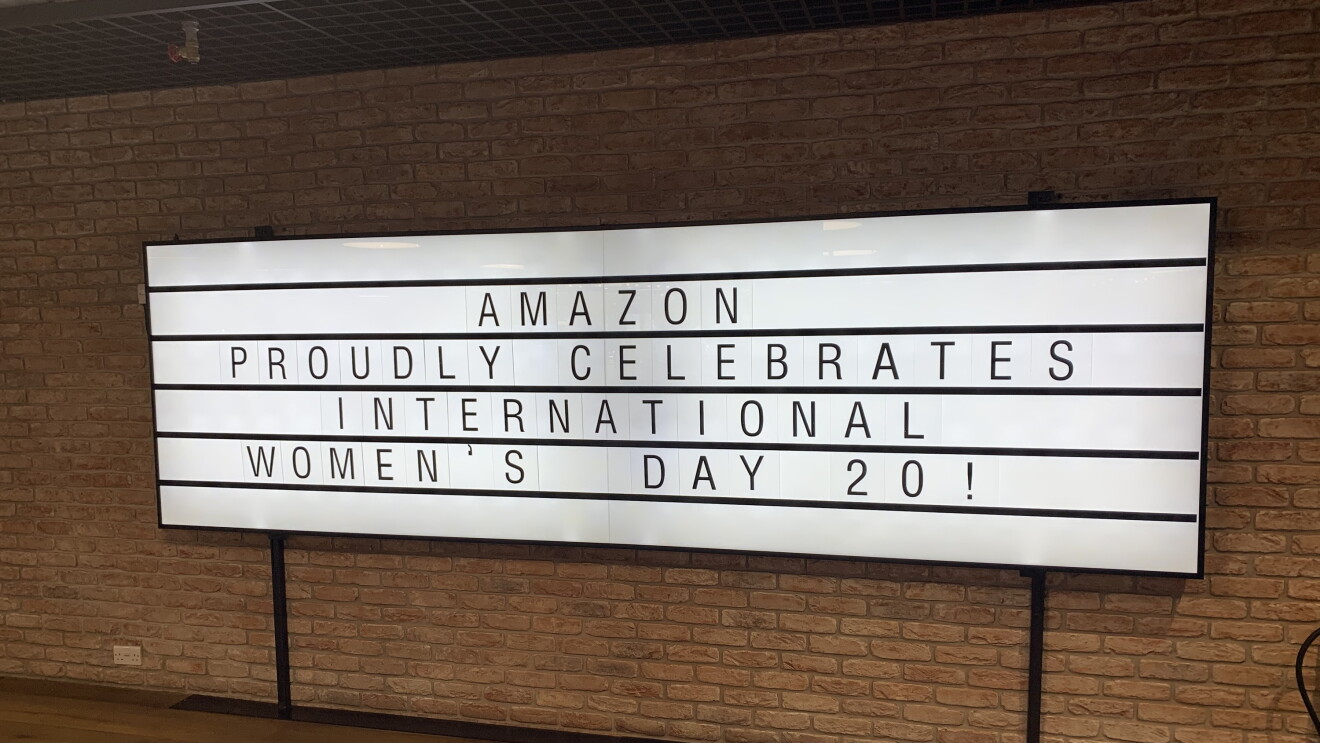 A large lightbox sign at the Amazon offices reading "AMAZON PROUDLY CELEBRATES INTERNATIONAL WOMEN'S DAY 20!". The sign is hanging on a brick wall. 