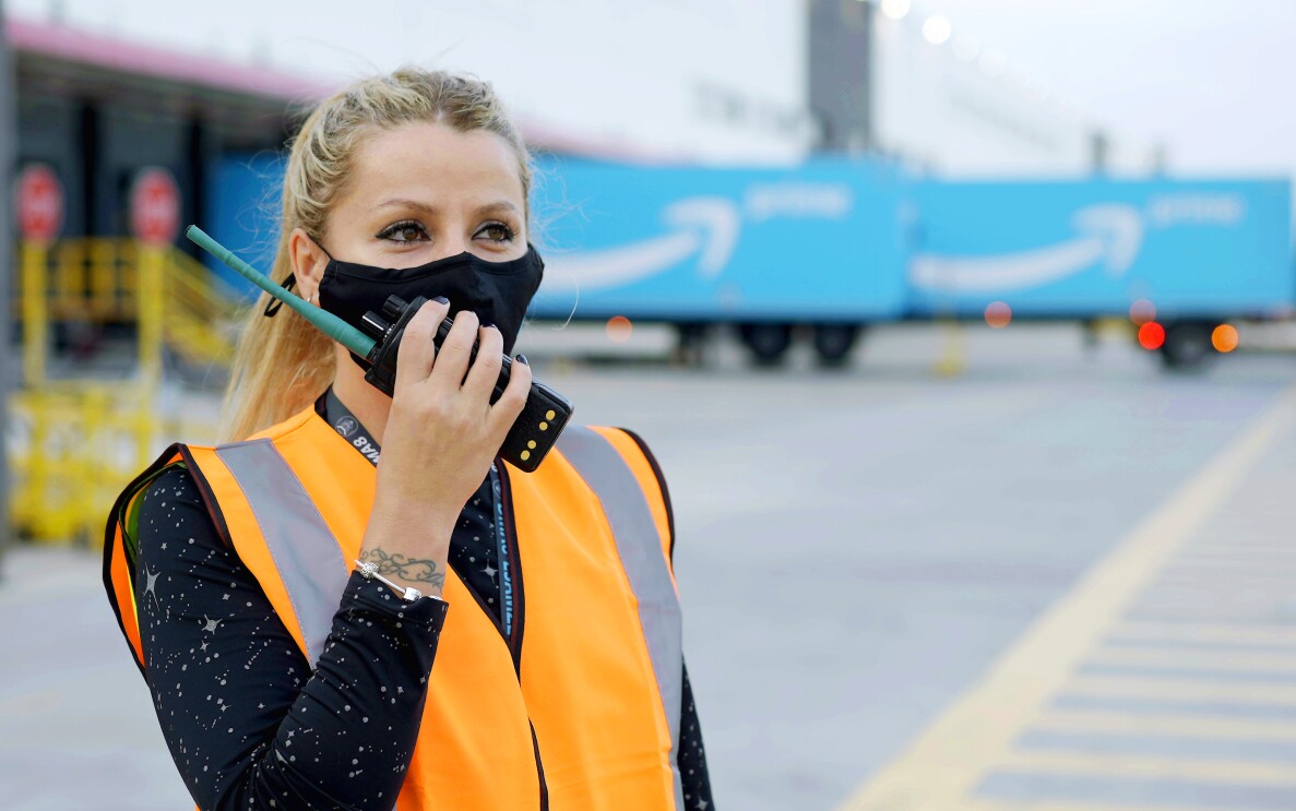 Ramona Moraru, supervisora de operaciones en la estación logística de Getafe. En el exterior del centro logístico con dos camiones de Amazon Prime de color azul con la sonrisa de Amazon en blanco. Ramona está hablando por un walkie talkie. Tiene el pelo claro, recogido en una cola, Con la mano derecha agarra el walkie talkie, tiene las uñas pintadas de negro. Lleva un tatuaje en la muñeca izquierda y un pulsera. Los ojos oscuros y maquillados y una mascarilla negra de Amazon. Lleva un chaleco de color naranja y un jersey negro con estrellas de color gris.