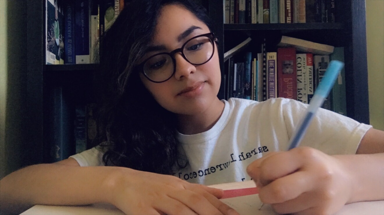 An image of a young woman writing at her desk. She is wearing glasses and a white t-shirt.