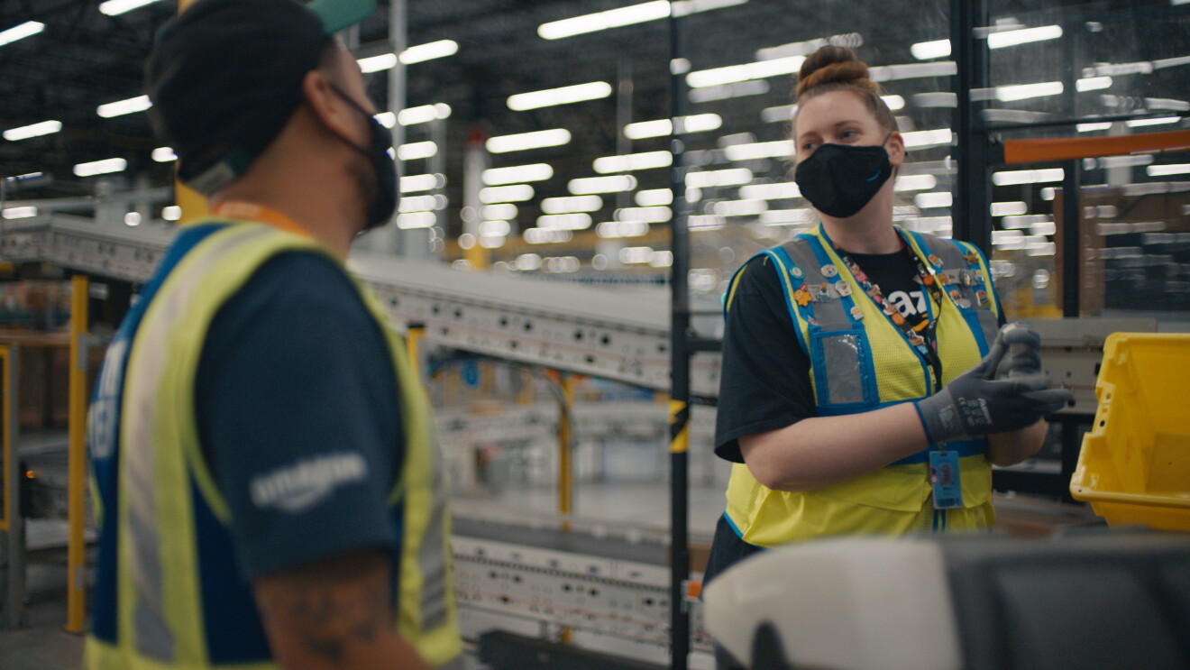 An image of a woman talking to her coworker while working in an Amazon fulfillment center. Both are wearing black masks with the Amazon logo on them and yellow safety vests.