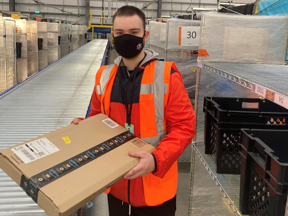 Finlay Shouler, Sortation Associate at Amazon's delivery station in Sheffield , pictured holding an Amazon parcel next to a conveyor belt.