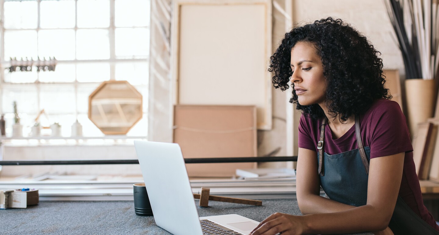 Un'imprenditrice di fronte al suo laptop in una bottega sta guardano lo schermo. Indossa un grembiule da lavoro, ha i capelli ricci.