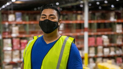 A male Amazon employee wearing a face mask and safety vest looks at the camera as he stands on a platform above the floor of a fulfillment center.