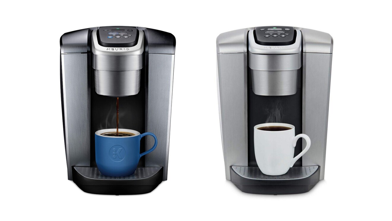 An image of two Keurig Coffee makers side by side. One is darker grey and the other is a lighter silver. The dark grey machine has a blue mug in it with coffee pouring into the mug. The silver machine has a white mug in it with coffee being poured into it.