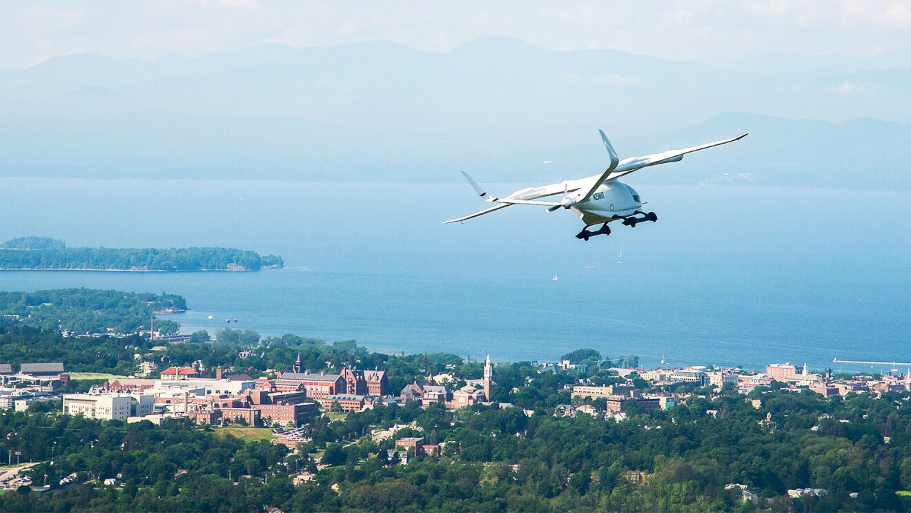 Small airfcraft flying over tree-filled land that is dotted with buildings, and a body of water beyond it