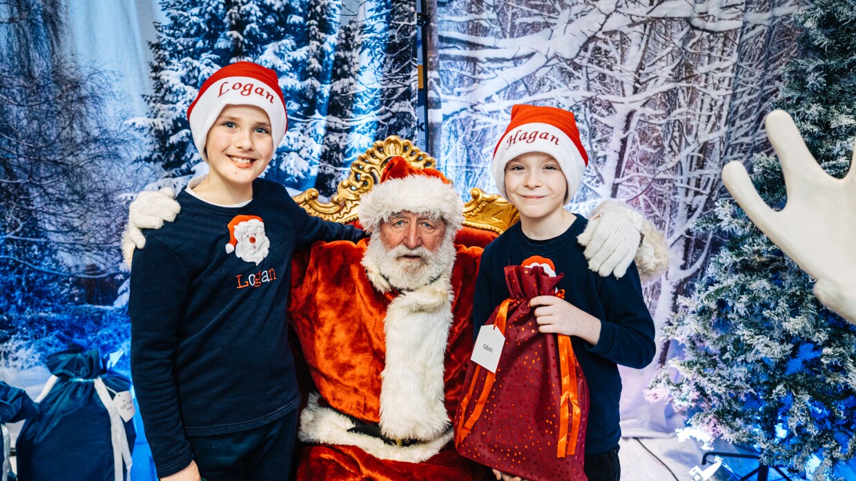 Two Children standing either side of Santa Claus in Santa's Grotto. They are each wearing Christmas hats.