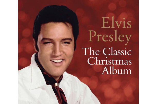 Elvis Presley wears a white button down and burgundy neck kerchief with a gold clasp. Behind him, we see out-of-focus lighting against a red background.