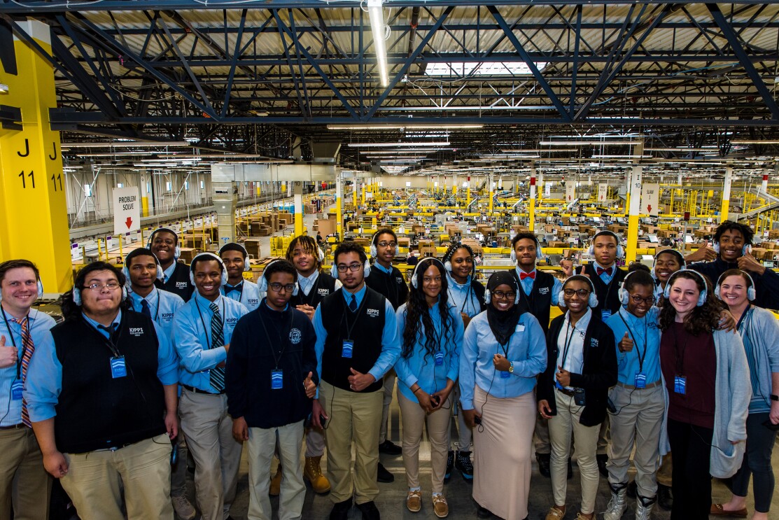 High school students wearing school uniforms pose as a group in an Amazon fulfillment center while taking a tour of the facility.