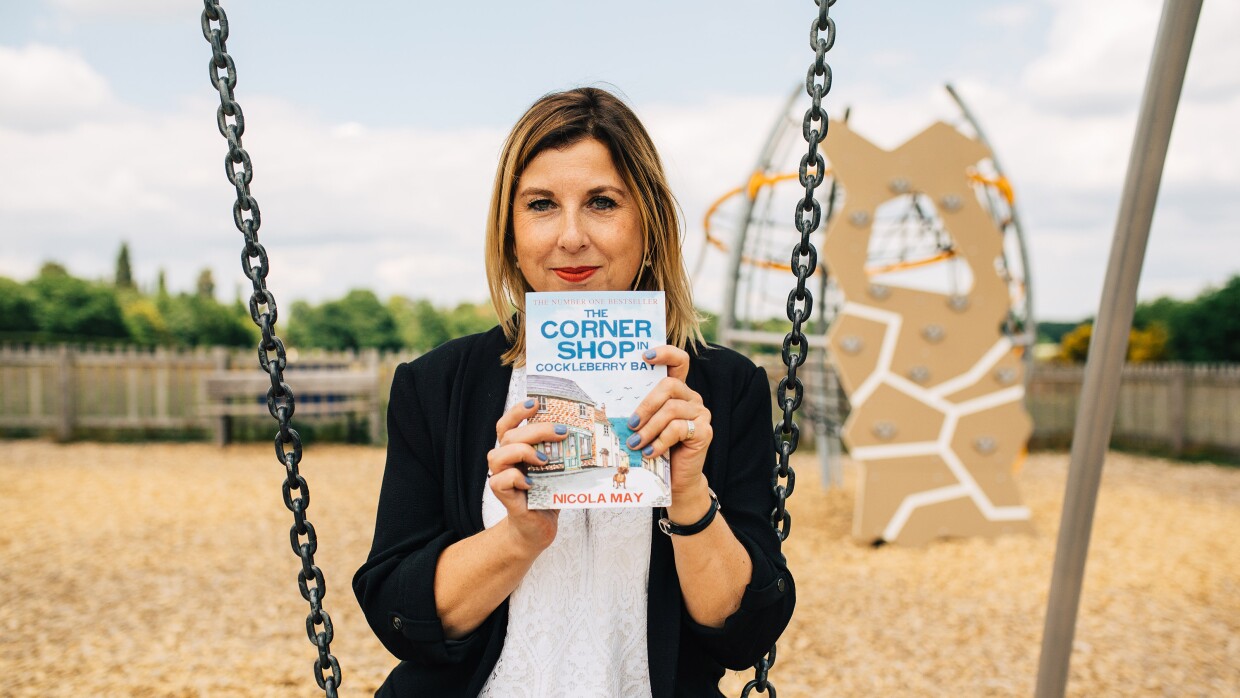 KDP author Nicola May sitting on a swing, holding here newest novel: The Corner Shop in Cockleberry Bay