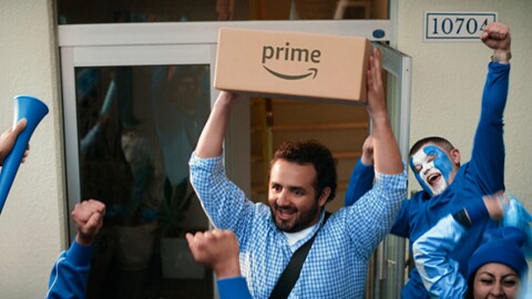 An image of a man holding a box with the Amazon Prime logo with a crowd of people wearing blue and white cheering him.