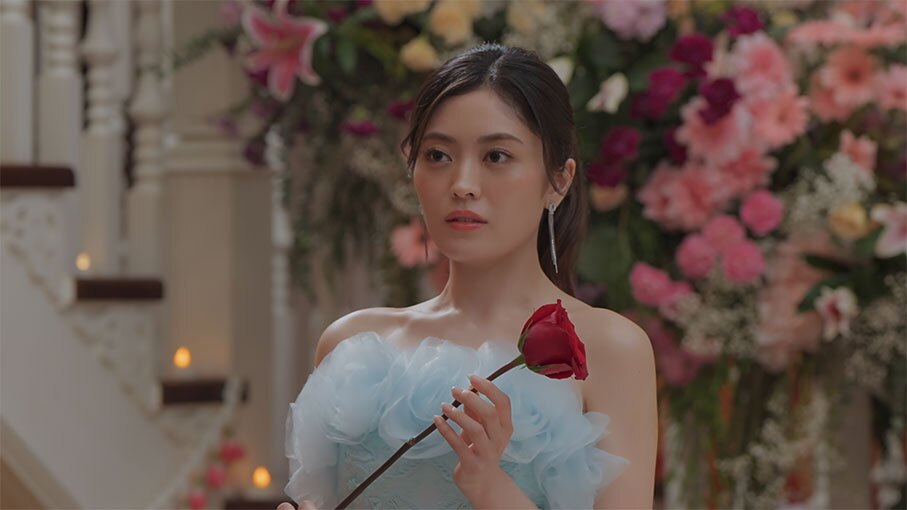 たくさんの生花が飾られた屋内に立つ女性。薄い水色のドレスを着用、体の前で一輪の赤いバラを持っている。