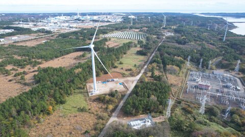 木々や畑の中に風力発電用の風車が複数立っている様子を写した空撮写真。右側には発電所の施設。